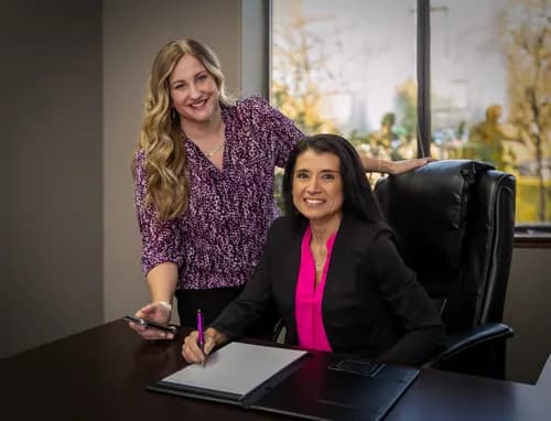 Two smiling people at an office desk, one seated with a pen and notebook and one standing beside the chair