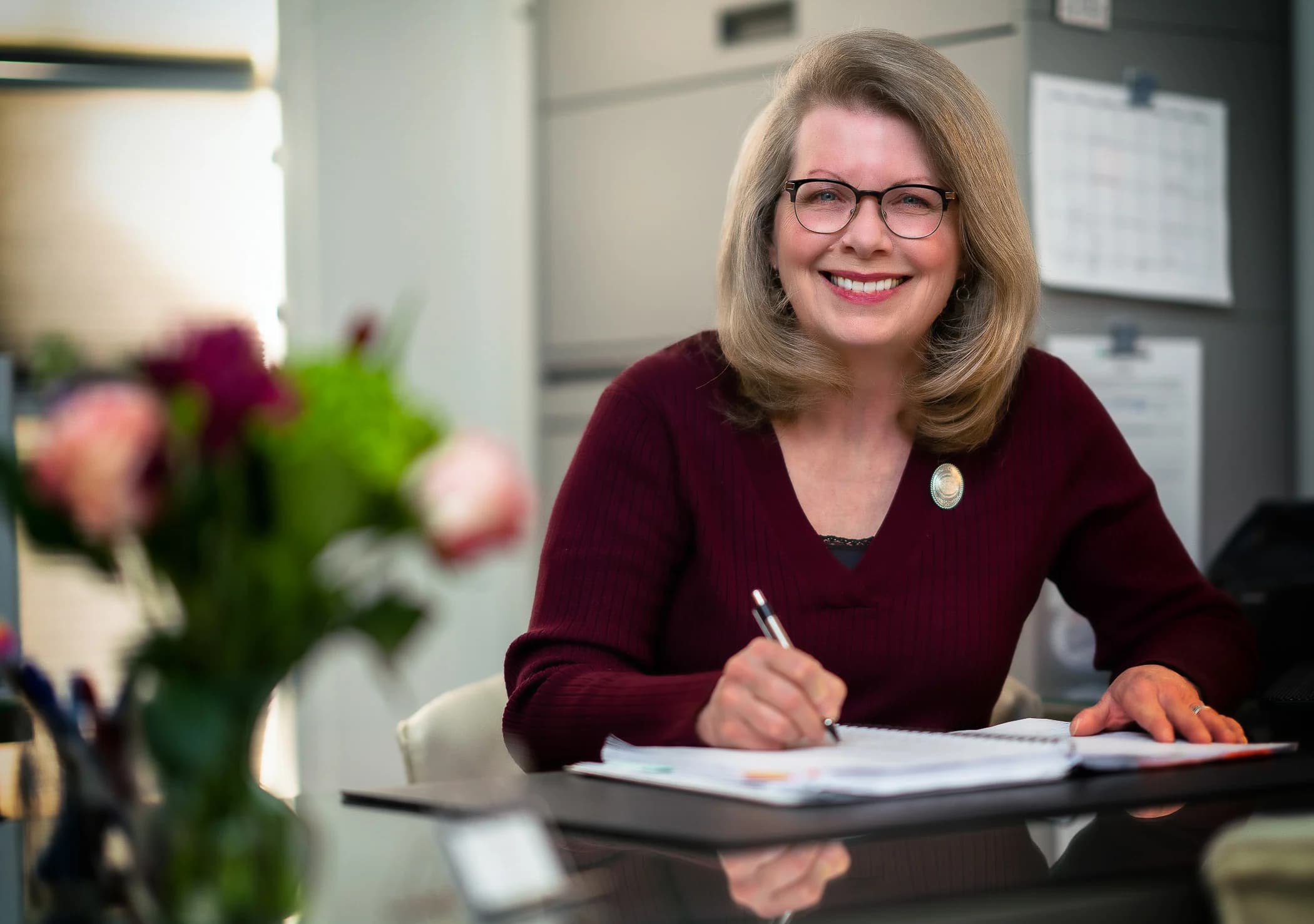 Smiling person with glasses in a maroon sweater writing at a desk with flowers in the foreground