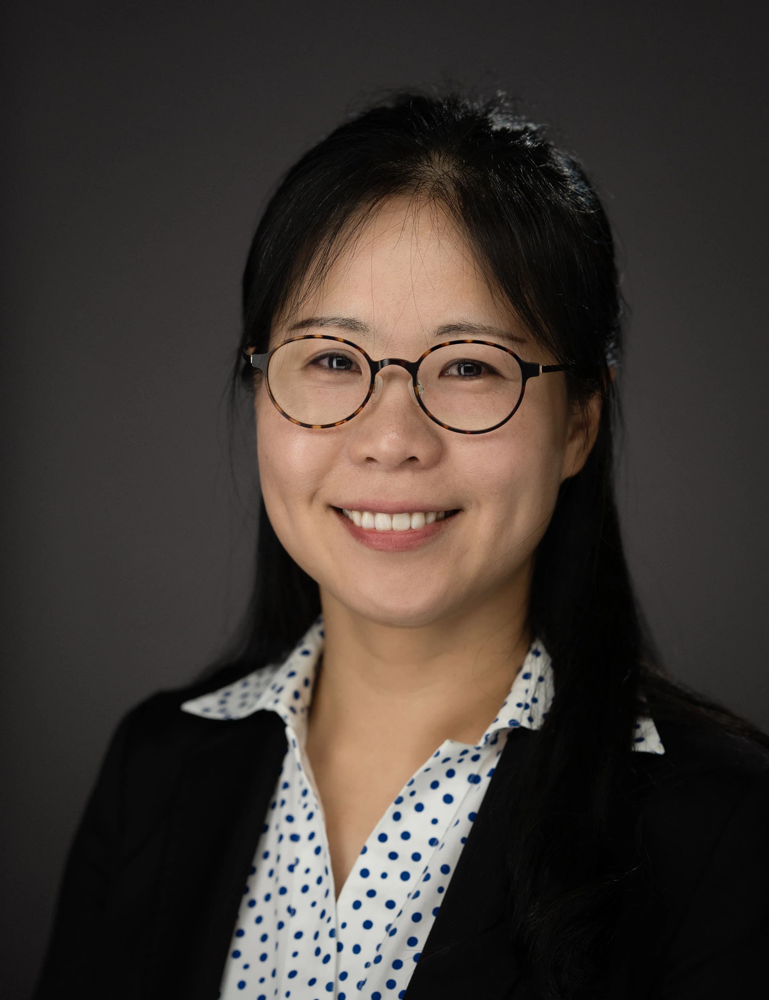 Headshot of a smiling person with long black hair and round glasses in a black blazer and polka-dot blouse